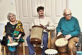 Three adults seated indoors playing hand drums during a UAA cultural session.